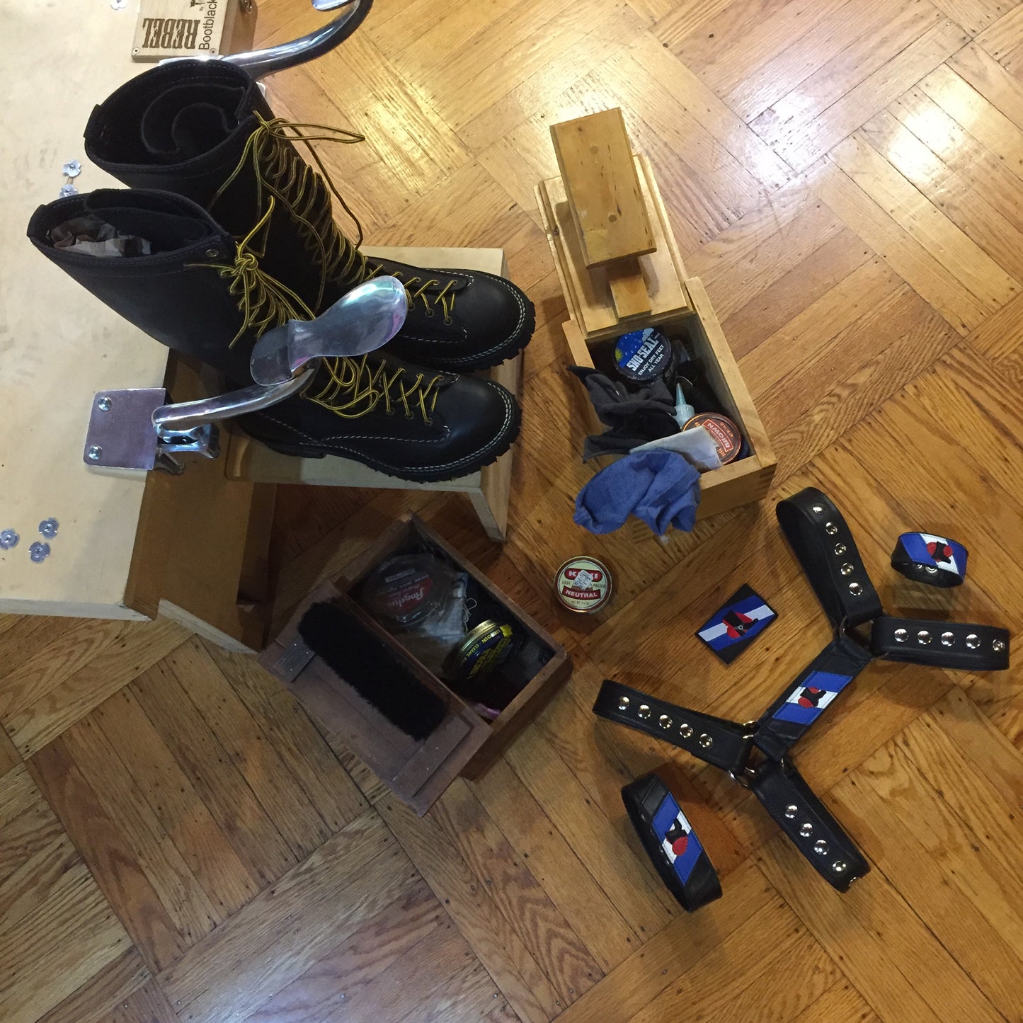 A display of bootblack materials along with the bootblack pride accessories; wrist cuff, leather patch, arm band and harness flag attached to a harness.